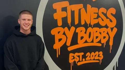 Bobby is on the left of the picture, dressed from head-to-toe in black. He is standing in front of the logo of his gym 'Fitness by Bobby est 2023' which is written in orange.