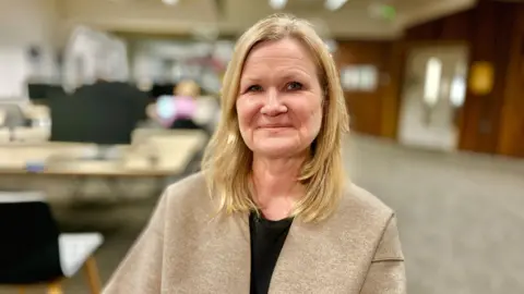 Martin Giles/BBC Cathrine Jansson-Boyd smiles at the camera. She has blonde hair that reaches her shoulders. She wears a beige coat and a black top. A blurred teaching area is visible behind her. 