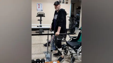 Jared Hughes A man in a black cap, t-shirt and hoodie. He has two prosthetic legs and is standing up and holding his weight on two parallel metal bars on either side of him. Behind him is a wheelchair