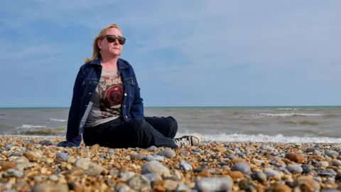 Jim Holden / BBC A woman with blonde shoulder -length hair who wears sunglasses that sits on a pebble beach near the water edge. The sea is behind her and the sky is blue. She wears a blue denim jacket and a top with a large red rose. 