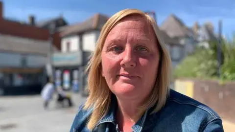 Stacey Brady in denim jacket sitting outside in sunny weather. She has shoulder-length blonde hair. It is a head and shoulders shot of her.