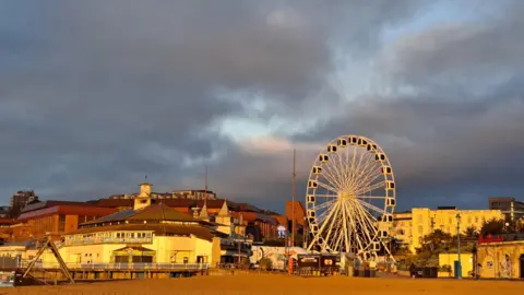 EvaDorset Dark moody skies create a backdrop for the early yellow sunshine illuminating the white seafront buildings and Ferris wheel at Bournemouth Pier with the beach in the foreground.