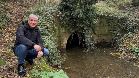 Justin Guy Peter Williams in front of the Clackers Brook culvert. He is crouching by a low-level waterway passing through a tunnel and smiling. 