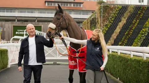 Anthony Devlin Paul is smiling at the camera at a racecourse alongside a brown race horse wearing a red coat. A woman with long blonde hair is holding the horse's reigns and smiling at Paul. There are two hedges either side of them and a white gate behind them. 