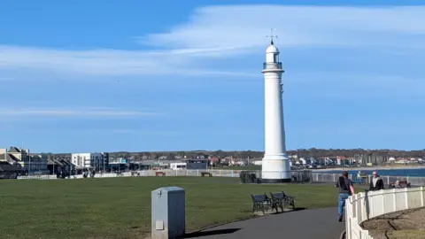 Neal Nimbus A white lighthouse overlooks the sea in Sunderland. There is a green manicured park on the left and buildings from the city in the background.