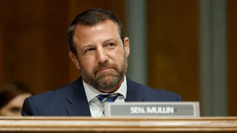 Mullin looks straight ahead and slightly smiles as he sits in a navy suit behind a placard of his name