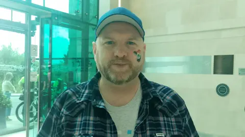 Scott Cuthbertson. He has a beard, wearing a blue cap, blue check shirt and grey t-shirt. He has a rainbow heard on his cheek.