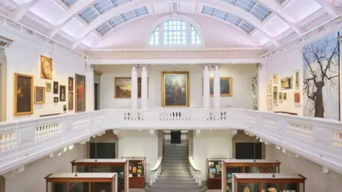 A large white room that has two floors. The upper floor is filled with large paintings of different sizes. The ground floor has a large staircase with large cabinets with artefacts inside. 