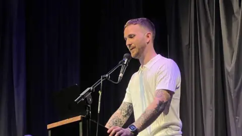 Andrew Taylor stands at a lectern with a microphone in front of him. He wears a white polo shirt and a watch. He has short sandy hair, a short beard and tattooed arms.