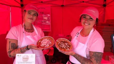 North East Lincolnshire Council Two women holding large pieces of cookie dough with toppings on. They are both wearing pink caps, pink T-shirts and pink aprons. They are stood inside a tent, smiling. 