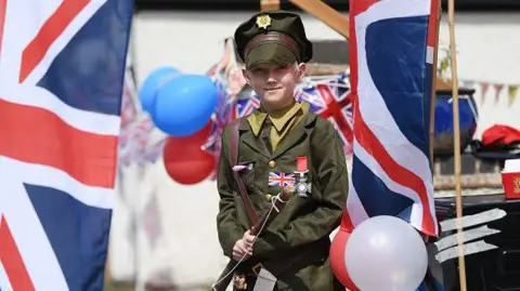 Getty Images A boy poses in military fancy dress during celebrations for VE Day in 2020 for the 75th anniversary. He is wearing a uniform with a flag on his jacket and he is carrying a satchel. there are Union flags around him and red, white and blue balloons.