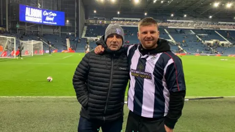 Aidan Smith Two men are standing beside a football pitch, one has his arm around the other. The man on the left is wearing a black puffa jacket and a beenie hat with a West Bromwich Albion logo on, the man on the right is smiling and has a long sleeved West Bromwich Albion top on. 