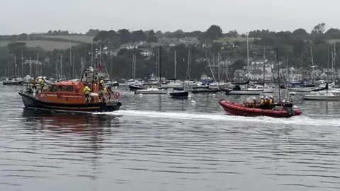 Two RNLI lifeboats head out to sea from Falmouth Harbour. Dozens of yachts are anchored in the background.