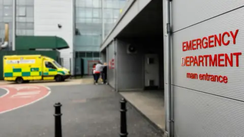 Getty Images The outside of a building with "Emergency Department main entrance" on the wall. An ambulance is parked outside and some people are standing at the door.