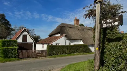 Nigel Wass This picture shows a white rendered thatched roof cottage under a blue sky. At the forefront of the photo is a wooden sign, leaning to the right with the words in white reading "To the church" with a white hand pointing to the right.