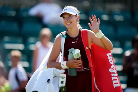 Katie Boulter with her bag and racket over her shoulder waving and smiling