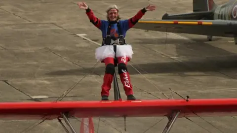 A grey haired woman wearing a parachute harness and tutu stands on the top wing of a red biplane with arms outstretched on an airfield.