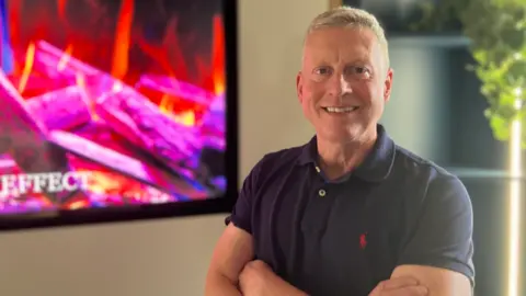 A man in a blue polo shirt looks at the camera, smiling. He has short grey hair, and his arms are crossed. To the side of him is a screen displaying a picture of a fire
