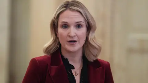 Helen McEntee has shoulder length blonde hair and is wearing a dark red blazer, with a black blouse underneath 