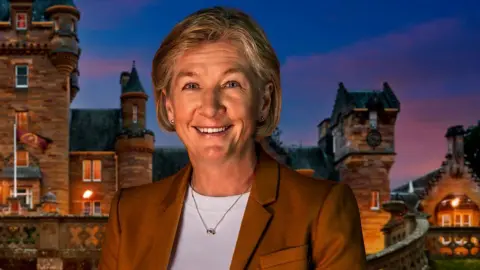 BBC A woman wearing a brown blazer and white top. She is stood in front of a castle at sunset.