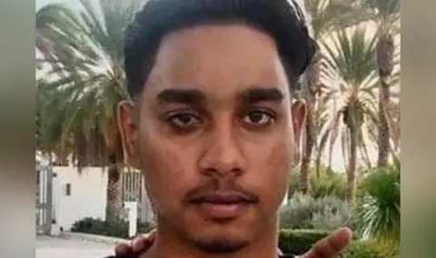 Family Handout Close-up of a young man's face. He has dark hair and eyebrows and a light mustache and goatee. He is outside, surrounded by palm trees and low bushes. A white wall and gate can be seen in the background.