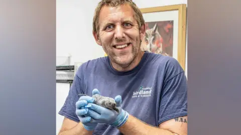 Birdland Park and Gardens Alistair King smiles at the camera and holds up the small, grey, king penguin chick. It is about the same size of the palm of his hand.
