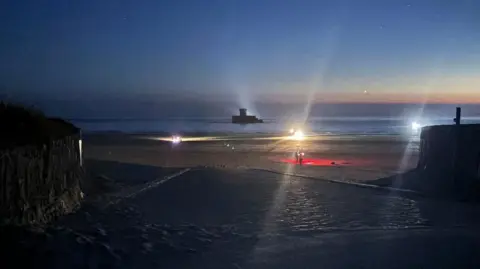 A beach at night time. The sky is dark blue with some orange. There are bright lights on the beach. There is a person walking along the beach with a red light. 