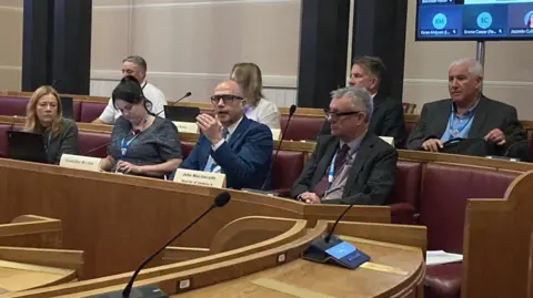Councillors sit in a council hall for a meeting. Two women and two men sit on the front row, with three men and one woman behind them.