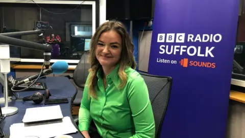 Vikki Irwin/ BBC Jenny Riddell-Carpenter is sitting in the BBC Radio Suffolk studios. She has long blonde hair and is smiling at the camera She is wearing a bright green shirt. 