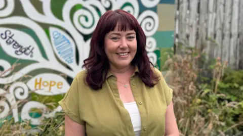 Siobhan McKinley is a young woman with shoulder-length hair which has been dyed a dark read colour. She is smiling at the camera. She is wearing a green dress with a white shirt underneath and a silver necklace. She is standing in front of some bushes and a large mural which has strength, hope and community written on the trees.