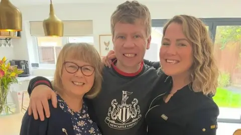 HAYLEY JANCESKI A woman wearing glasses, a navy floral dress and a blue cardigan, a man wearing a grey Newcastle United sweatshirt and a woman wearing a blue jacket stand next to each other in a kitchen. They have their arms around each other's shoulders and are all smiling at the camera.