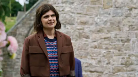 A woman stands with her hands in her pockets in front of a brick wall.