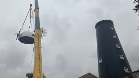 BBC A yellow crane is lifting a white wooden mill cap back onto the top of a tall black mill. the sky is grey and cloudy