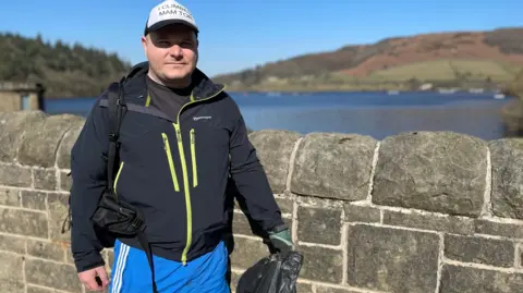 Shaun Broughton regularly goes to Ladybower Reservoir to litter pick. He is pictured wearing a cap and black coat with yellow flashes.