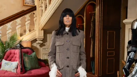 BBC Claudia Winkleman in the Traitors castle, wearing a brown jacket and white shirt with huge cuffs.