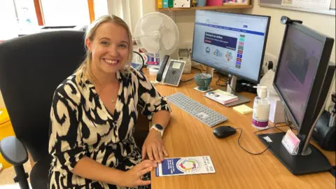 A female doctor with blonde hair sits at her desk, in her consulting room, next to her computer. She has a leaflet in front of her about wellbeing and social prescribing.