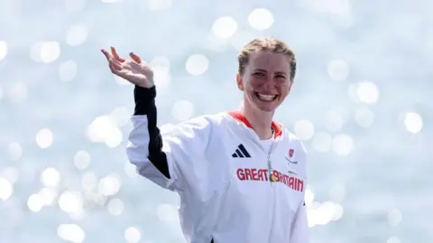 A woman wearing a white 'Great Britain' tracksuit smiling and waving at the camera. 