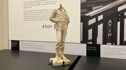 A plaster statuette about 3ft tall in yellowy white with a missing head and hand. It is sat on top of a black case.