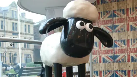 The original black and white Shaun the Sheep animation has been created into a sculpture. It has been positioned on a platform in front of a large glass window and a wall featuring Union Jack flag wallpaper. 