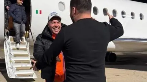 A man embraces another man after getting off a small passenger jet. The man facing the camera is smiling. He's wearing a black jacket, orange tshirt and black jacket.