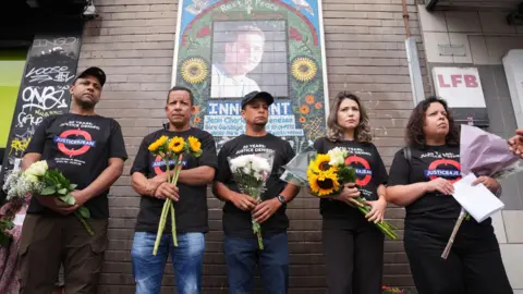 PA Media A group of people stand in front of a memorial mural of Jean Charles de Menezes, each holding flowers. They wear matching black T-shirts reading “20 Years: Justice Denied – Justice4Jean” and appear solemn and reflective.