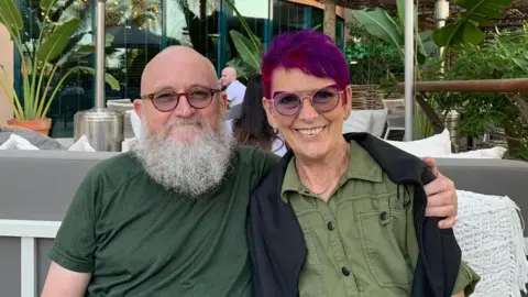 Cancer Research UK A man with a bald head, large white beard and glasses holds his arm around a woman with purple hair and glasses. The pair are smiling and are sitting on a chair outside. 