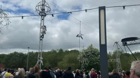 An acrobat performs high up while hanging down from a wire. Crowds below watch her.