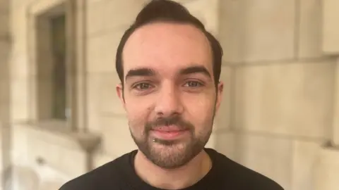 A man looks directly at the camera, he has dark hair, and a dark beard. He is wearing a black t shirt. 