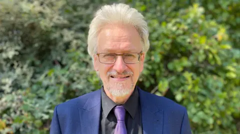 A man with grey hair and a goatee and glasses smiles looking into the camera. He's wearing a blue jacket, dark shirt and a purple tie. Behind him is a green bush, slightly out of focus. It is a head and shoulders shot of him.