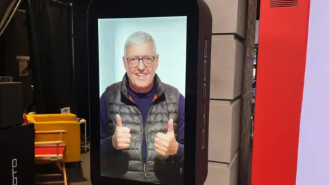 Proto Hologram Picture of hologram version of Bill on a large screen, wearing a purple jumper and black gilet. He smiles and gives two thumbs up.