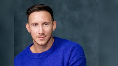 Rhys Batten A headshot of Rhys Batten, looking at the camera in front of a dark background. He is wearing a blue jumper.