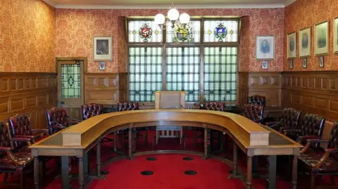 MANX SCENES The Legislative Council chamber, which has brown wooden panelling on the walls and features a horseshoe-shaped table with maroon leather chairs around it on a red carpet. There is a stained-glass window at the back of the chamber with various crests on it. 