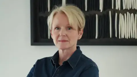 Helen Molyneux Helen has short cropped blonde hair and is looking at the camera. She has a dark blue denim jacket on. It is a head and shoulders shot of her. She is in front of a white wall with a black and white framed painting behind her. 