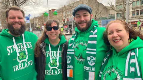 Two men and two women pose for a photo in Dublin ahead of the St Patrick's Day parade through the city.  The men both have beards and the women both have long hair.  They are all wearing bright green hooded tops. One of the women is wearing black sunglasses with two shamrocks above her head. 